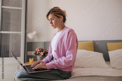 Calm young caucasian girl with blonde hair uses laptop sitting on bed indoor. Serious woman wears casual clothes at home. Concept working mood.