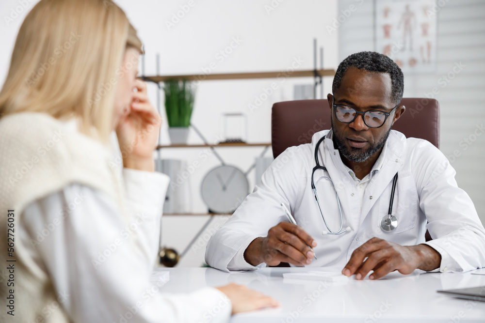 Fototapeta premium African American doctor, receives a patient, listens to complaints, makes diagnostics. Writes data. Doctor's appointment.