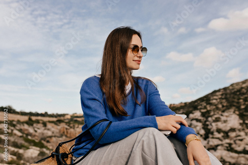 European cute woman with dark hair in sunglasses wearing blue sweater enjoying travelling among mountains in sunny day. Travel girl enjoying vacation 