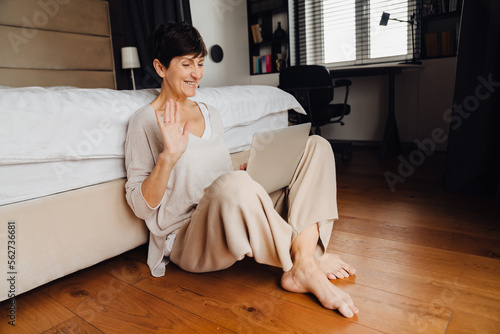 Smiling mature woman making video call via laptop and waving hand while sitting in bedroom