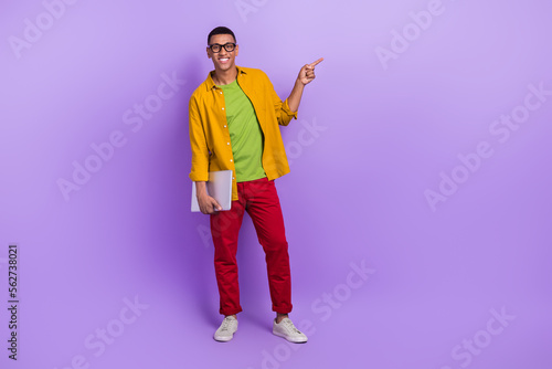 Full body portrait of cheerful handsome guy hold netbook direct finger empty space isolated on purple color background © deagreez