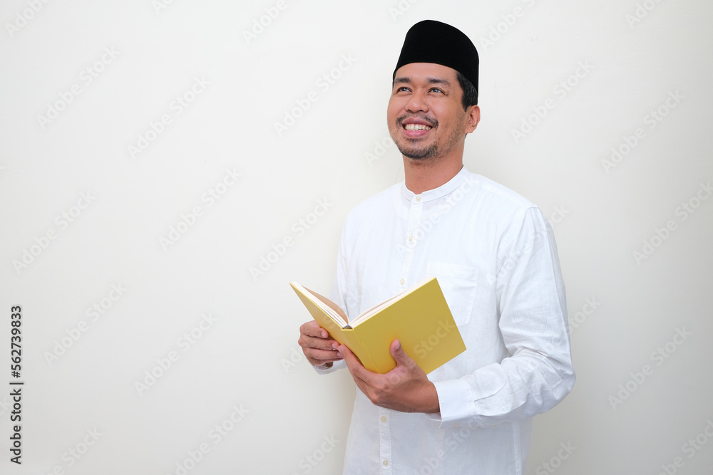 Moslem Asian man smiling and looking above while holding a book Moslem Asian man smiling and looking above while holding a book