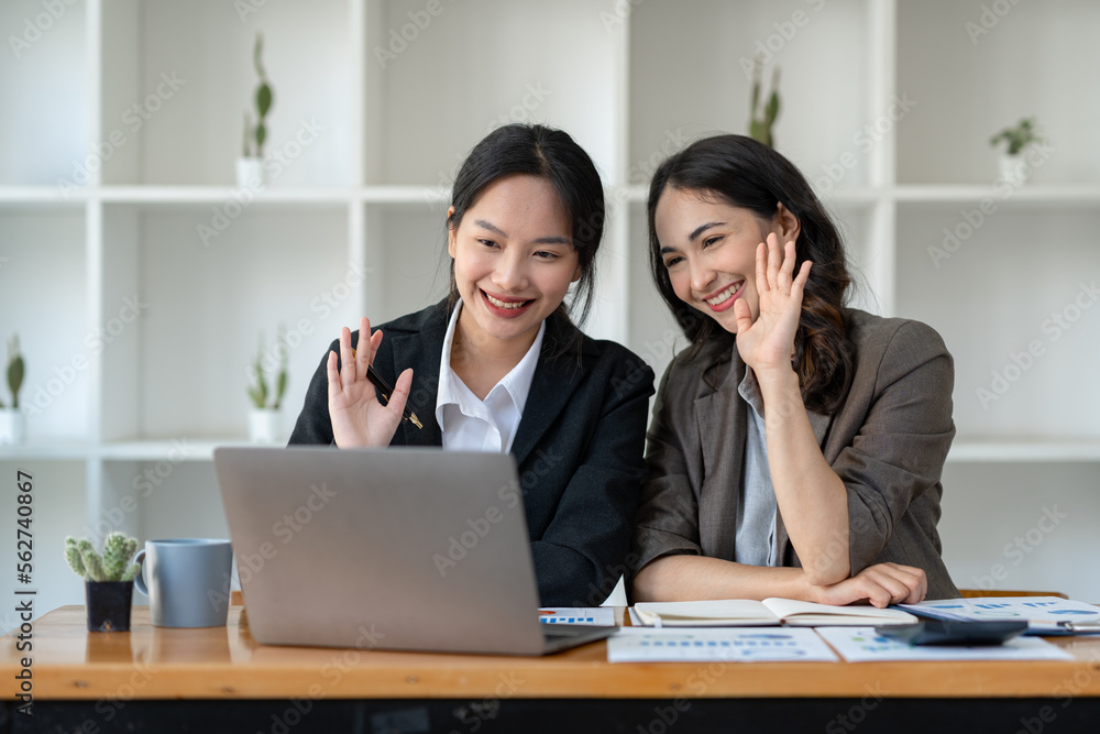 Image of two beautiful Asian businesswomen waving greeting online through a laptop at the table with a calculator, graph, and chart showing business growth data at the office. Image of two beautiful Asian businesswomen waving greeting online through a laptop at the table with a calculator, graph, and chart showing business growth data at the office.