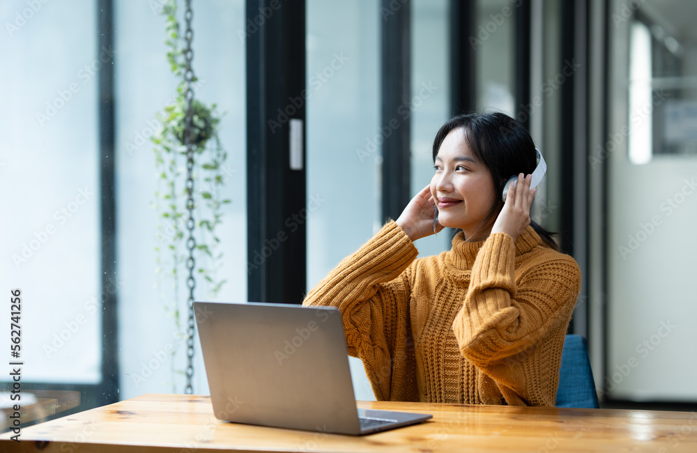Fototapeta premium Young Asian women enjoy listening to music wearing headphones and feeling relaxed by stretching their arms. Count to the beat at her desk.
