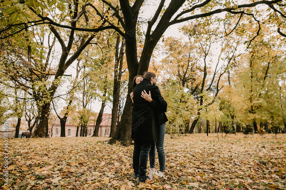 Side view of adult man and woman hugging during meet in autumn park. Couple of man and woman in love, dressed in casual apparel, cuddling while standing near tree in forest