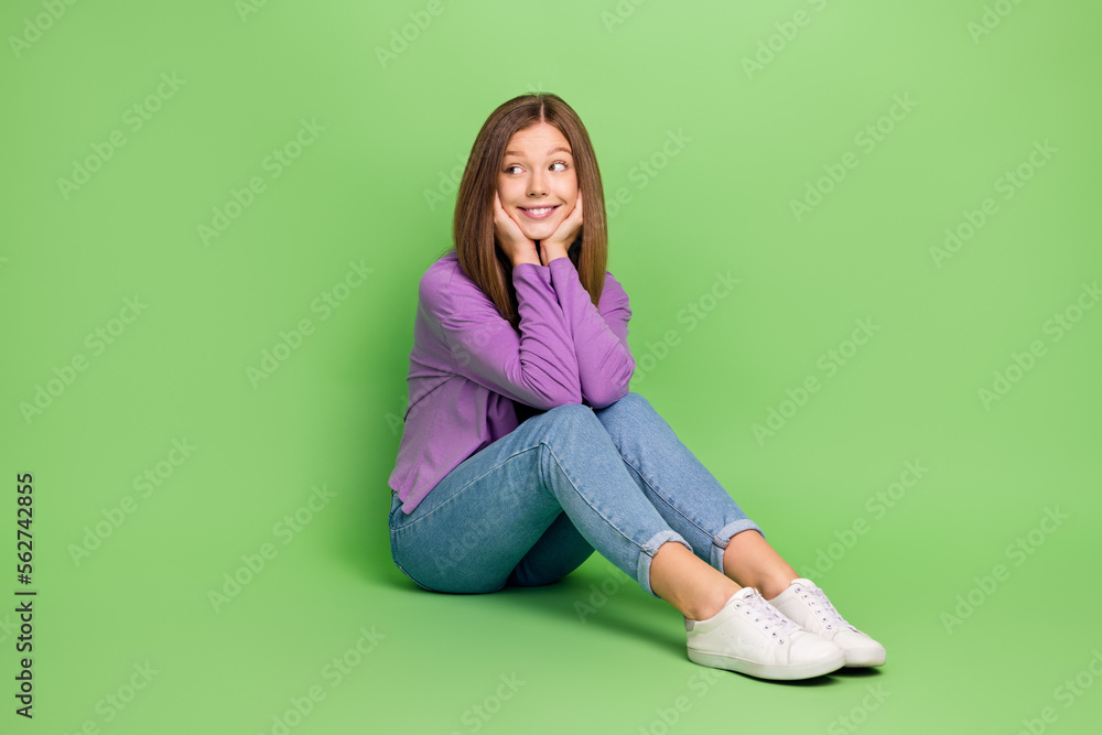 Full length photo of good mood girl with straight hairdo wear purple pullover sitting look empty space isolated on green color background Full length photo of good mood girl with straight hairdo wear purple pullover sitting look empty space isolated on green color background
