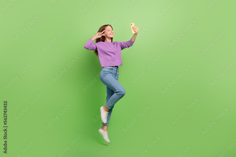 Full size photo of cute positive girl dressed jeans purple pullover hold smartphone showing v-sign isolated on green color background Full size photo of cute positive girl dressed jeans purple pullover hold smartphone showing v-sign isolated on green color background