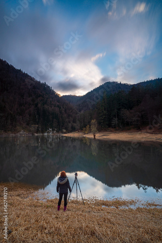 View of solo female photographer with a tripot on a lake side on mountains wirh reflction on water and colorful sunset clouds on sky
