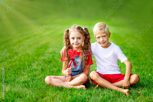 children girl and boy blonde with a big lollipop heart in the summer on the lawn on the green grass, the concept of the valentine's day holiday, space for text