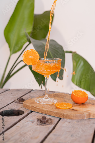 Sok drink pomarańczowy plusk. Nalewanie drinka, przygotowanie drinka. Szkło do drinka, kieliszek. Sok drink w kieliszku na nodze. Egzotyczny drink, egzotyczne zielone liście w tle. Cocktail Aperol.