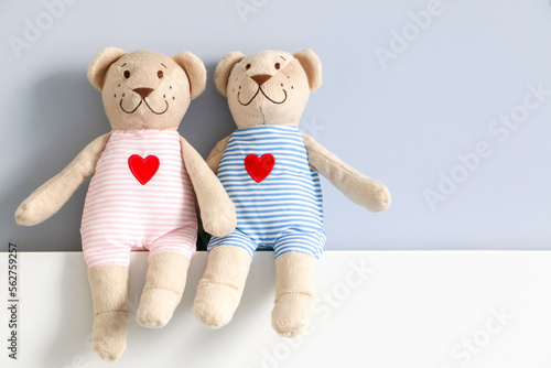 A couple of his and hers teddy bear in light pink and blue striped clothing with red heart pattern, sitting on headboard of bed against light blue wall. Stuffed toy gift.