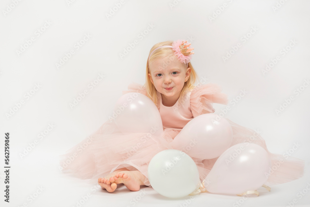 Smiling blonde girl in pink dress and hoop on white background. Isolate. Mischievous child sits on floor in balloons Smiling blonde girl in pink dress and hoop on white background. Isolate. Mischievous child sits on floor in balloons