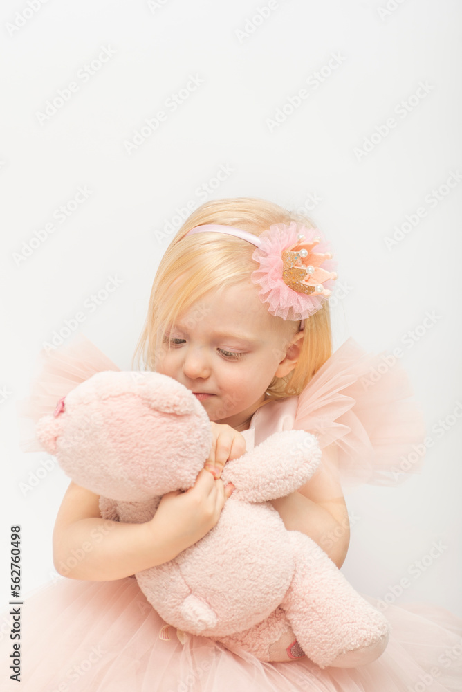 Pretty little girl hugs pink teddy bear. Blonde girl in lush pink dress and hoop decoration. White background. Vertical Pretty little girl hugs pink teddy bear. Blonde girl in lush pink dress and hoop decoration. White background. Vertical