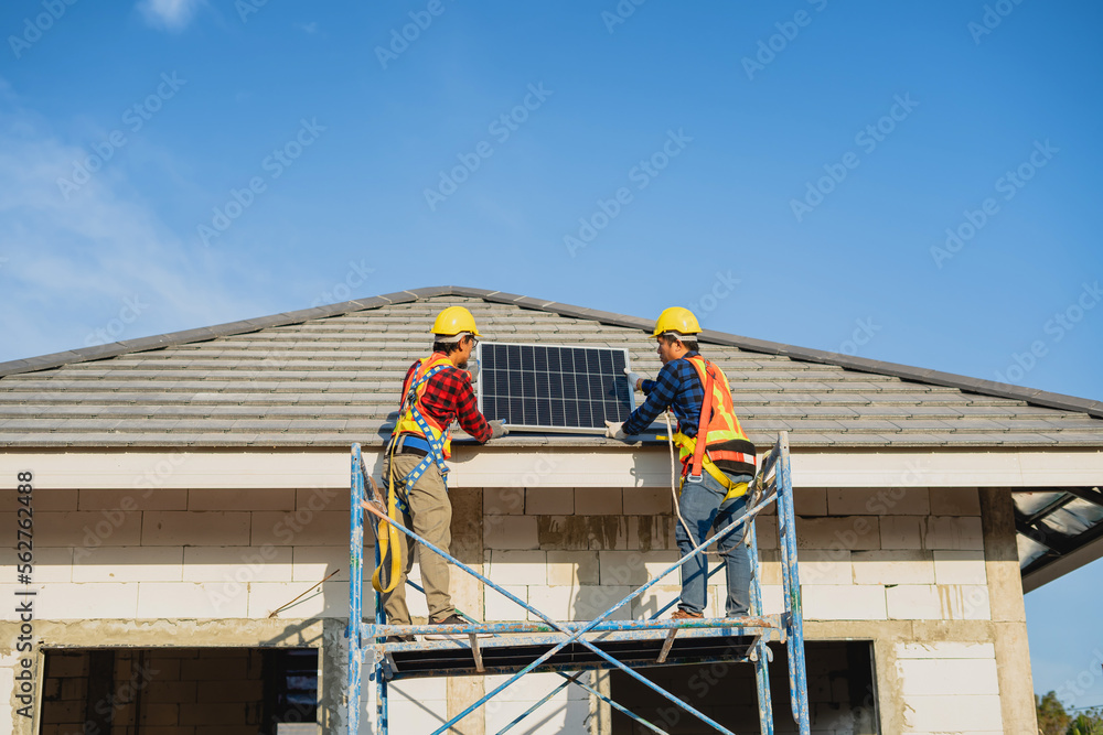 Two male construction workers in protective clothing hold solar panels on the roof of a house.