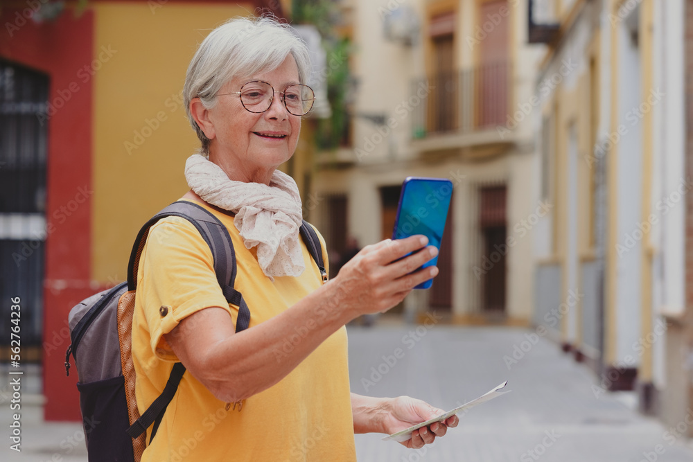 Obraz premium Carefree senior traveler woman carrying backpack visiting the old town of Seville looking at phone, smiling elderly lady enjoying travel and discovery