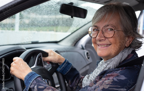 Senior woman driver driving her car on a rainy day. Elderly woman holding the steering wheel looking smiling at camera