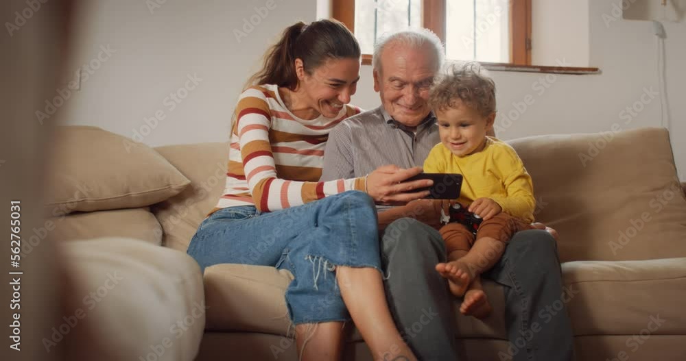 Happy Small Family Gathered Together, Sitting on a Sofa in the Living Room and Using a Smartphone. Mother, Son and Grandfather Watching Videos on a Mobile Phone and Laughing. They are Bonding Happy Small Family Gathered Together, Sitting on a Sofa in the Living Room and Using a Smartphone. Mother, Son and Grandfather Watching Videos on a Mobile Phone and Laughing. They are Bonding