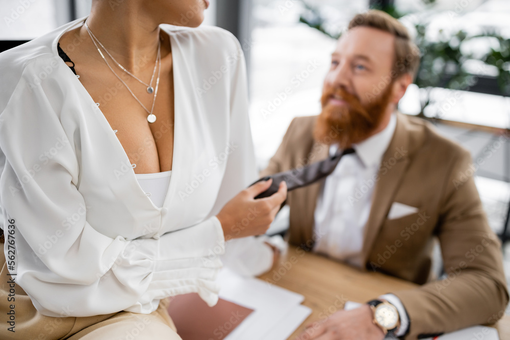 Obraz premium cropped view of african american woman with big breast sitting on desk and pulling tie of bearded coworker in office.