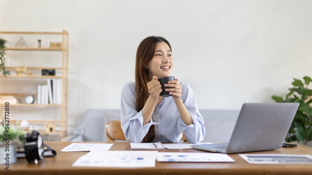 Beautiful Asian woman working on laptop and sipping coffee with smiling face in her home, Creating happiness at work with a smile, Freelancer working at home happily, Work from home. Beautiful Asian woman working on laptop and sipping coffee with smiling face in her home, Creating happiness at work with a smile, Freelancer working at home happily, Work from home.