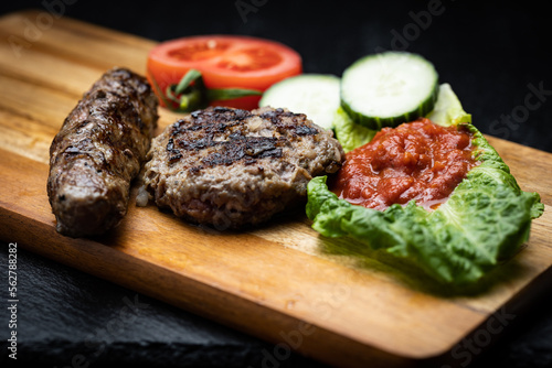Traditional Bulgarian homemade grilled ground meat sausages called kyufte and kebapche placed on wooden plank next to dipping sauce lyutenica and slices of cucumber and tomato