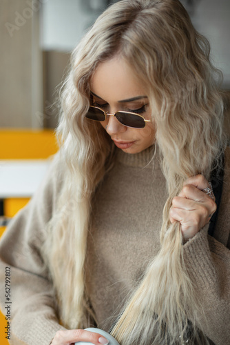 Fashionable beautiful girl with a blond curly hairstyle with vintage sunglasses in a beige sweater sits and combs her hair indoors