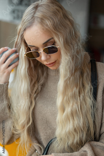 Fashion portrait of a beautiful stylish girl with a blond curly hairstyle with vintage sunglasses in a beige sweater