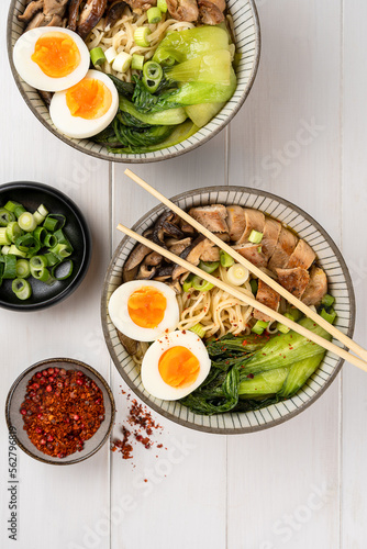 Two bowls of ramen soup with chicken breast, vegetables, mushrooms, pak choi and egg on a white wooden table and chopsticks. Top view, healthy eating concept