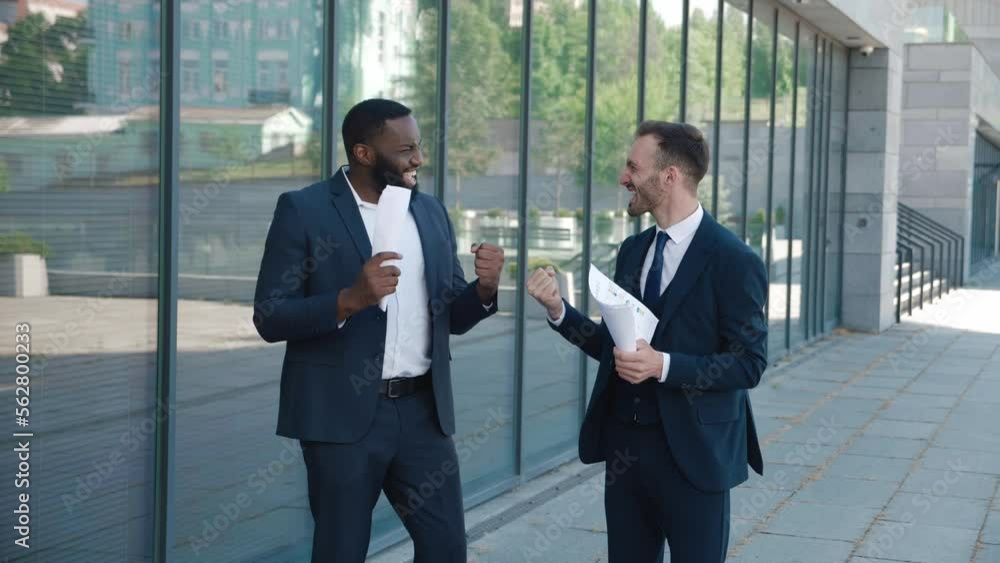 Two excited diverse business men in formal suits having a successful deal. Colleagues business men celebrating victory and winning, making yes gesture standing in city center near office building. Two excited diverse business men in formal suits having a successful deal. Colleagues business men celebrating victory and winning, making yes gesture standing in city center near office building.