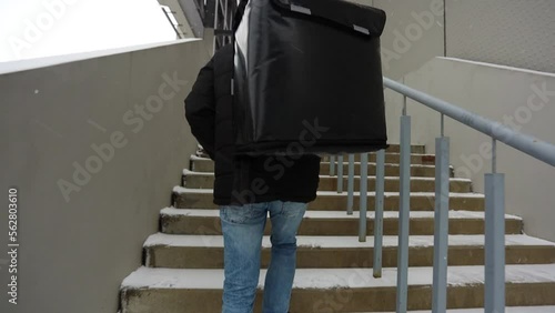 Courier with thermal backpack and delivering food. A man in a black jacket is walking down the stairs. The concept of the work of the delivery man