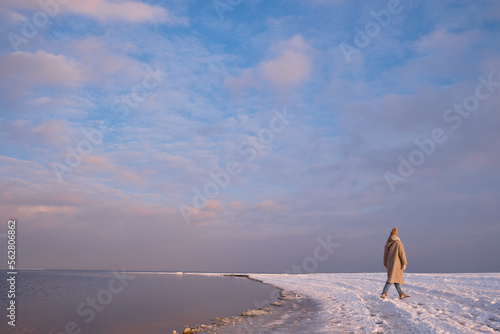 Stylish girl walks at sunset along the beach of the sea or ocean on a cool winter day. Travel concept. Walk on the sea or ocean. Amazing scenic outdoors view. Beautiful sunset on the seashore