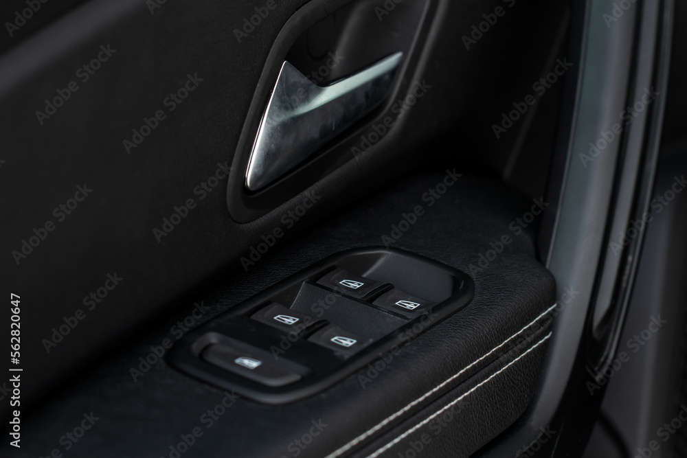 Car door handle with power window control. Dark leather interior of modern car. Dark black and orange car leather pleats stitch. Details of door handle with windows controls. Car window controls.