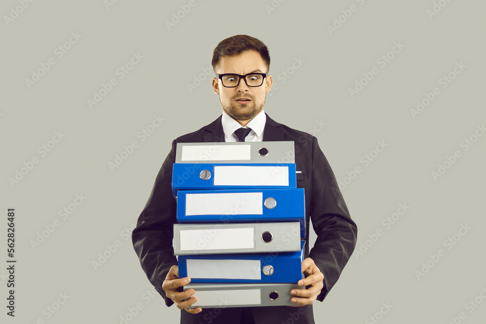 Bureaucracy and overworking concept. Angry office worker with pile of papers and documents is stressed by lot of work and deadlines. Man in suit angrily looks at stack of folders on gray background. Bureaucracy and overworking concept. Angry office worker with pile of papers and documents is stressed by lot of work and deadlines. Man in suit angrily looks at stack of folders on gray background.