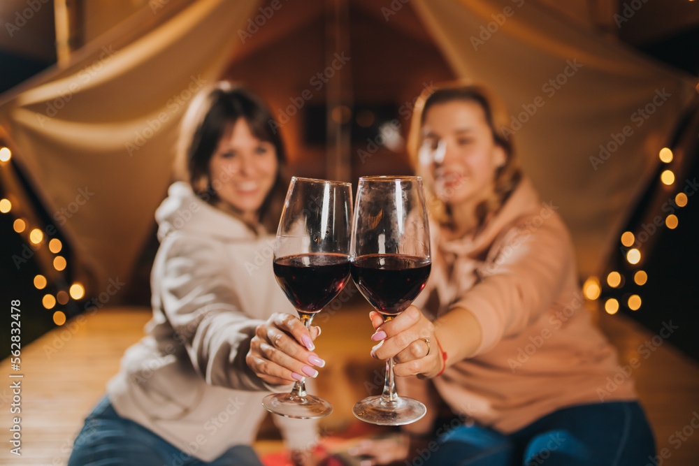 Two Smiling female friends drinking wine and eating fruits sitting in cozy glamping tent in autumn evening bonfire. Luxury camping tent for outdoor holiday and vacation. Lifestyle concept Two Smiling female friends drinking wine and eating fruits sitting in cozy glamping tent in autumn evening bonfire. Luxury camping tent for outdoor holiday and vacation. Lifestyle concept
