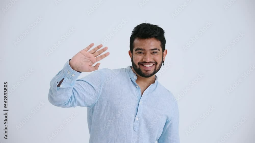 Smiling Indian man with piercing wearing blue shirt waving hand at the camera in the grey studio Smiling Indian man with piercing wearing blue shirt waving hand at the camera in the grey studio