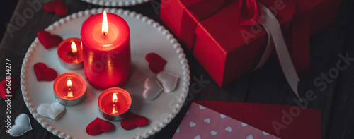Saint Valentine's Day celebration. Red burning candles, hearts, gift box, postcard on dark wooden background. Happy holiday. Table decor for festive dinner, romantic atmosphere. Banner photo