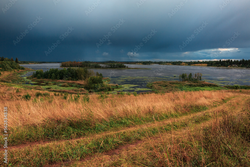 Landscape of a lake in a field, storm clouds, dirt road.