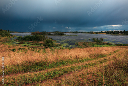 Landscape of a lake in a field, storm clouds, dirt road.