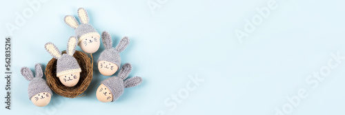 Five Easter eggs with funny faces and gray crocheted hats with bunny ears in a decorative nest of sisal on a blue background. Happy Easter concept. Banner