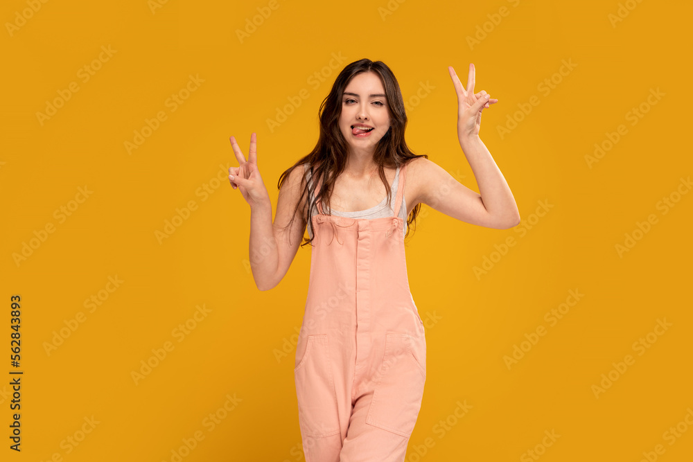 Positive, young, happy woman having fun in studio, sticking tongue and showing peace sign, victory gesture. Yellow background. Copy space. Positive, young, happy woman having fun in studio, sticking tongue and showing peace sign, victory gesture. Yellow background. Copy space.
