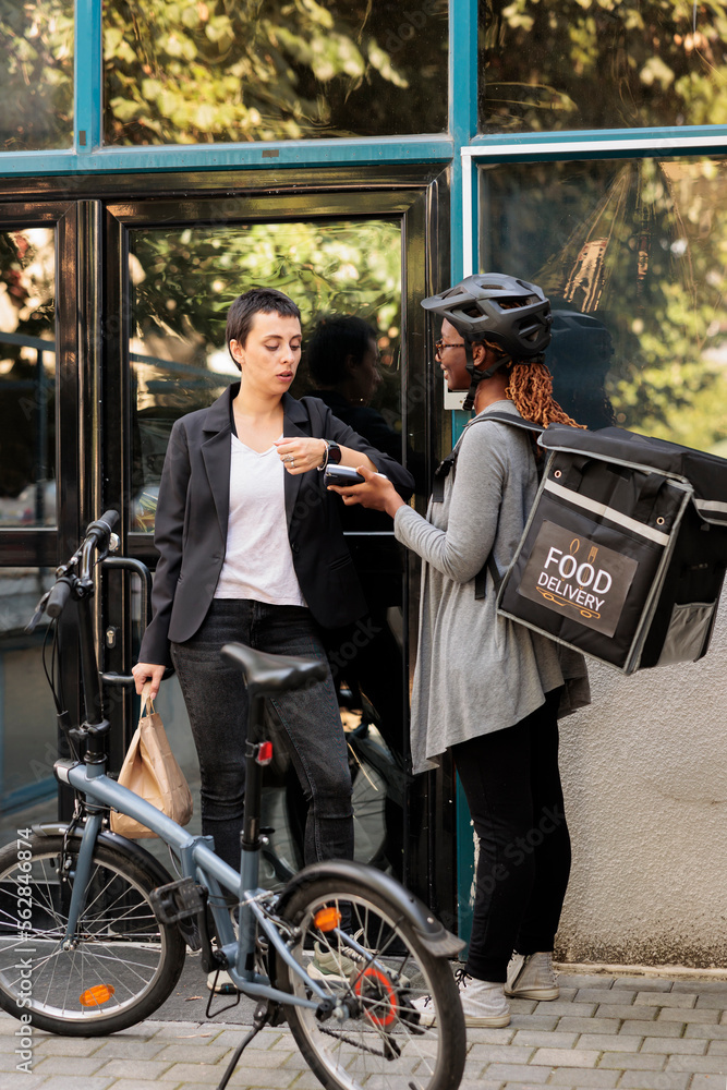 Naklejka premium Office employee paying for delivery with smart watch contactless payment transaction. African american courier holding pos terminal, standing near company building outdoors