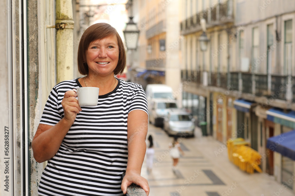 Middle ages woman holds cup of coffee or tea, standing on the balcony with Europe city street view in the morning. Mature female enjoying vacation in resort. Summer, weekend, tourism, travel