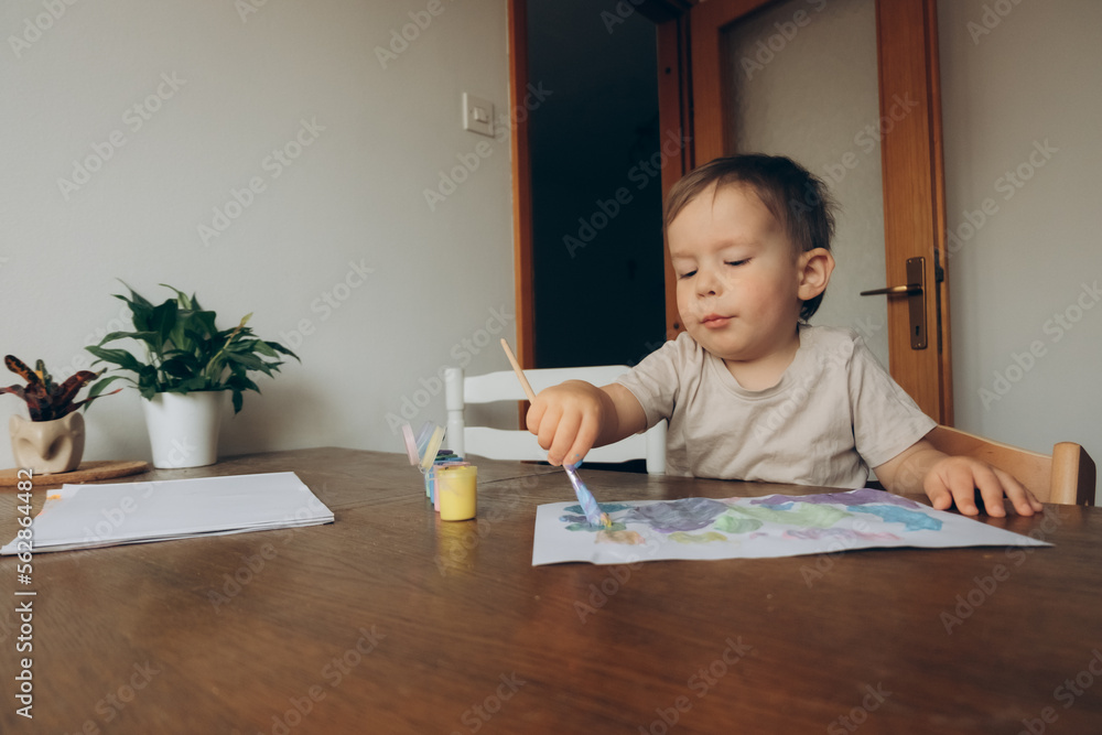 child draws with a brush and multicolored gouache sitting at the table drawing to his mother