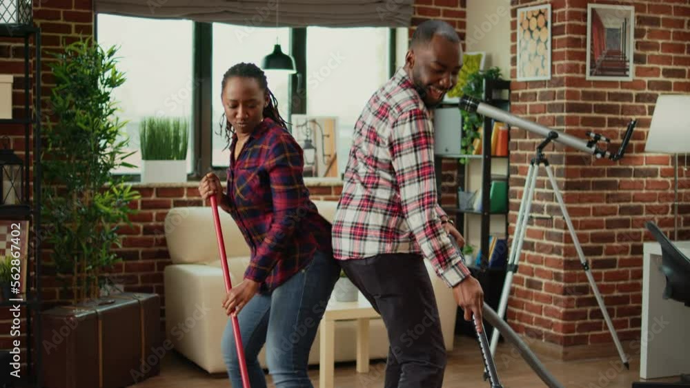 Cheerful people showing dance moves and doing spring cleaning, having fun with music while they clean wooden floors with mop and vacuum cleaner. Young couple smiling and doing chores. Cheerful people showing dance moves and doing spring cleaning, having fun with music while they clean wooden floors with mop and vacuum cleaner. Young couple smiling and doing chores.