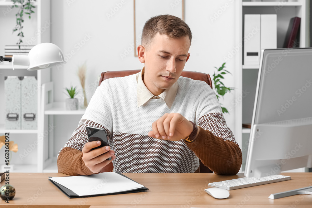 Young businessman with mobile phone looking at wristwatch in office