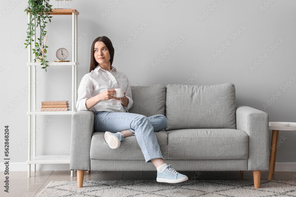 Fototapeta premium Young woman with cup of hot beverage sitting on sofa at home
