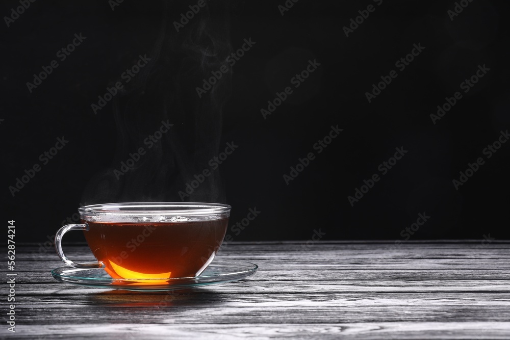 Fototapeta premium Glass cup of tea and saucer on wooden table against black background, space for text
