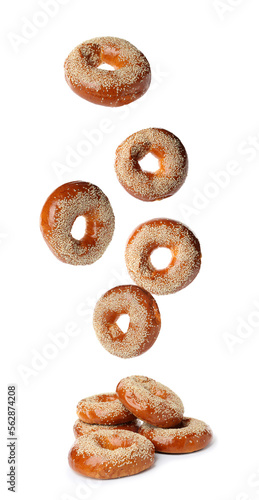 Many fresh bagels with sesame seeds falling into pile on white background