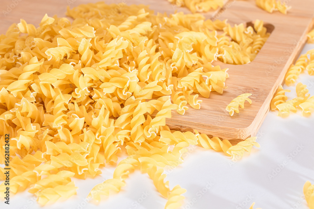 Fototapeta premium Uncooked Fusilli Pasta Scattered on Wooden Board and White Table. Raw and Dry Macaroni. Fat and Unhealthy Food