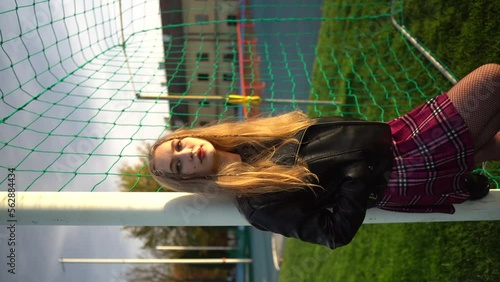 Blonde woman in skirt and lather jacket stands near soccer net at sport field