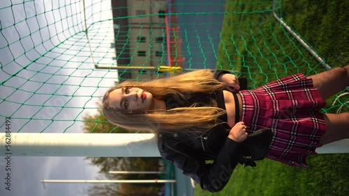 Blonde woman in skirt and lather jacket stands near soccer net at sport field